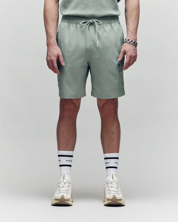 A person stands against a plain background wearing Psycho Bunny MENS RANDY BOARD SWIM SHORT - MB1000002 in sage green with cargo pockets, a coordinating shirt, white striped socks, cream sneakers, and a silver bracelet. The image is from mid-torso to feet.