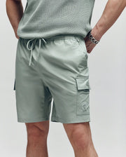 A person sports Psycho Bunny's MENS RANDY BOARD SWIM SHORT - MB1000002 in light sage green with cargo pockets and a drawstring waist, paired with a matching knit top and silver bracelet—ideal for a laid-back beach day.