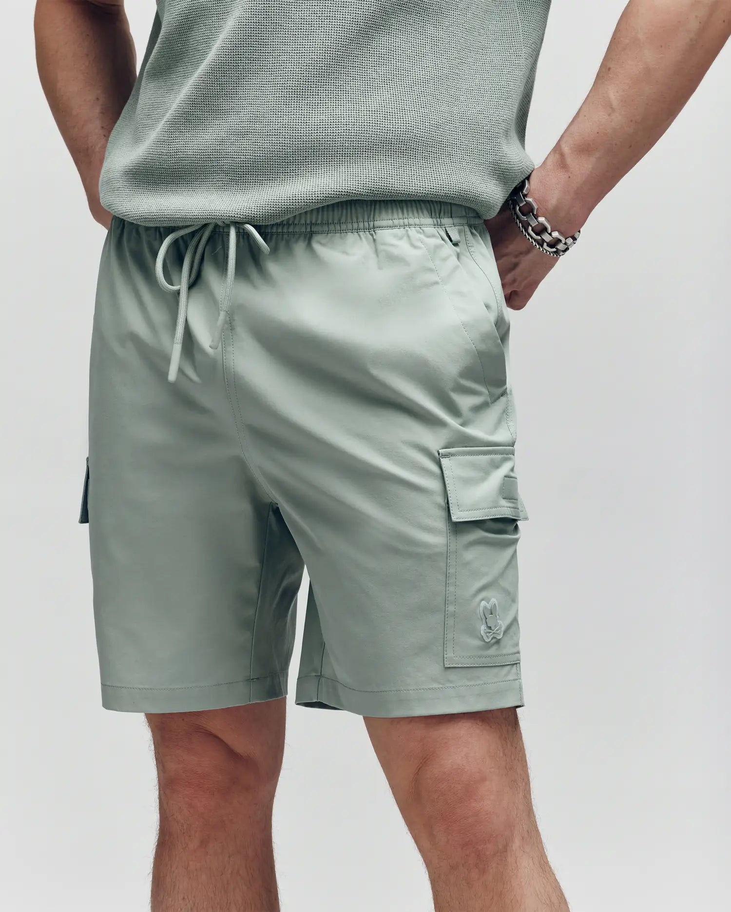 A person sports Psycho Bunny's MENS RANDY BOARD SWIM SHORT - MB1000002 in light sage green with cargo pockets and a drawstring waist, paired with a matching knit top and silver bracelet—ideal for a laid-back beach day.