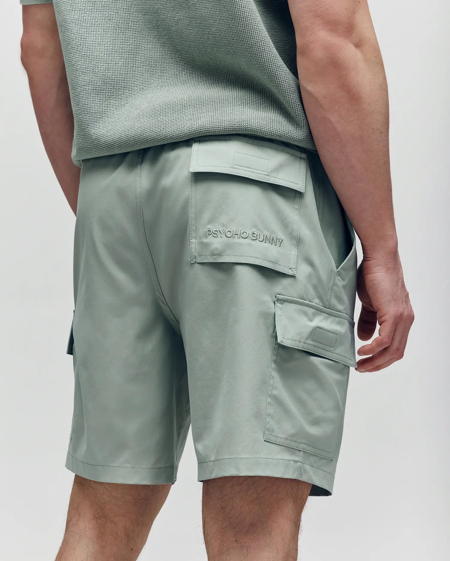 A person stands facing away in light green Psycho Bunny MENS RANDY BOARD SWIM SHORT - MB1000002 with cargo pockets and a matching textured shirt. "PSYCHO BUNNY" is embroidered on the back pocket, ideal beachwear.