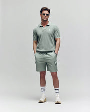 A man wearing sunglasses, a sage green polo, Psycho Bunny MENS RANDY BOARD SWIM SHORT - MB1000002 with cargo pockets, white socks with navy stripes, and white sneakers stands against a plain light background with hands in his pockets.