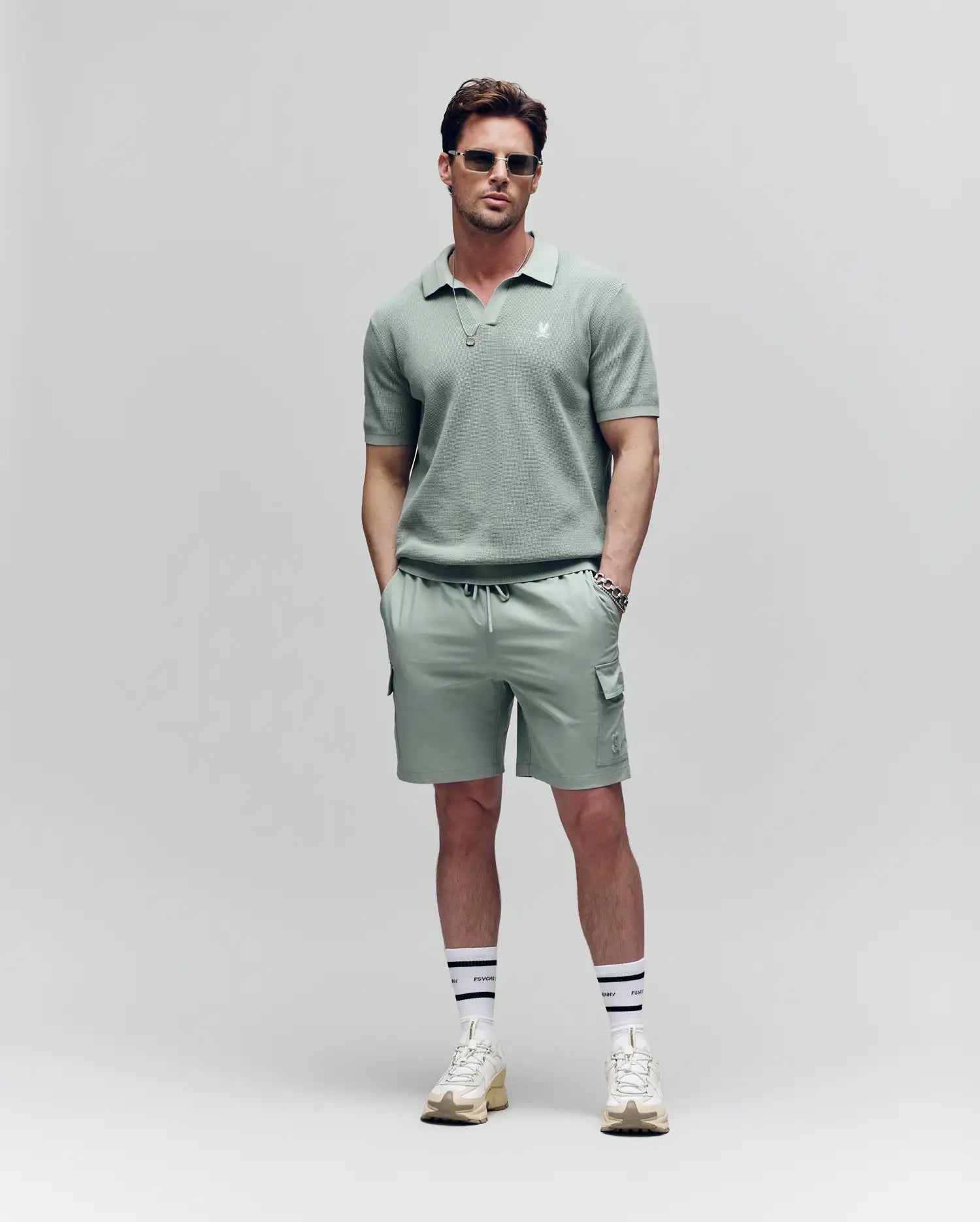 A man wearing sunglasses, a sage green polo, Psycho Bunny MENS RANDY BOARD SWIM SHORT - MB1000002 with cargo pockets, white socks with navy stripes, and white sneakers stands against a plain light background with hands in his pockets.