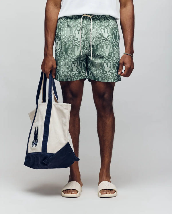 A person stands against a plain background wearing Psycho Bunny's MENS CHUCK ALL OVER PRINT LIGHTWEIGHT SWIM TRUNK (MB1000003) in green, a white t-shirt, beige slides, and holding a cream tote bag with navy accents and the Psycho Bunny logo.
