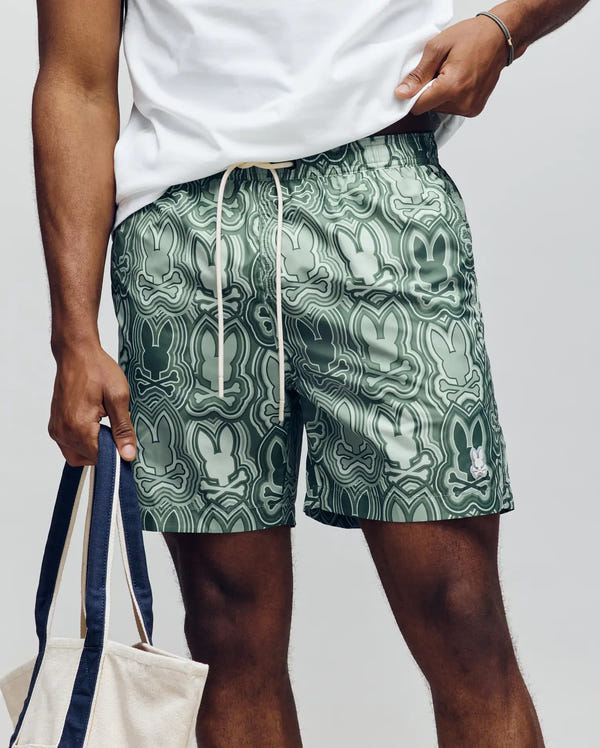 A person wears Psycho Bunny's MENS CHUCK ALL OVER PRINT LIGHTWEIGHT SWIM TRUNK MB1000003—green quick-dry recycled polyester shorts with abstract bunny faces—paired with a white shirt and holding a beige tote bag.