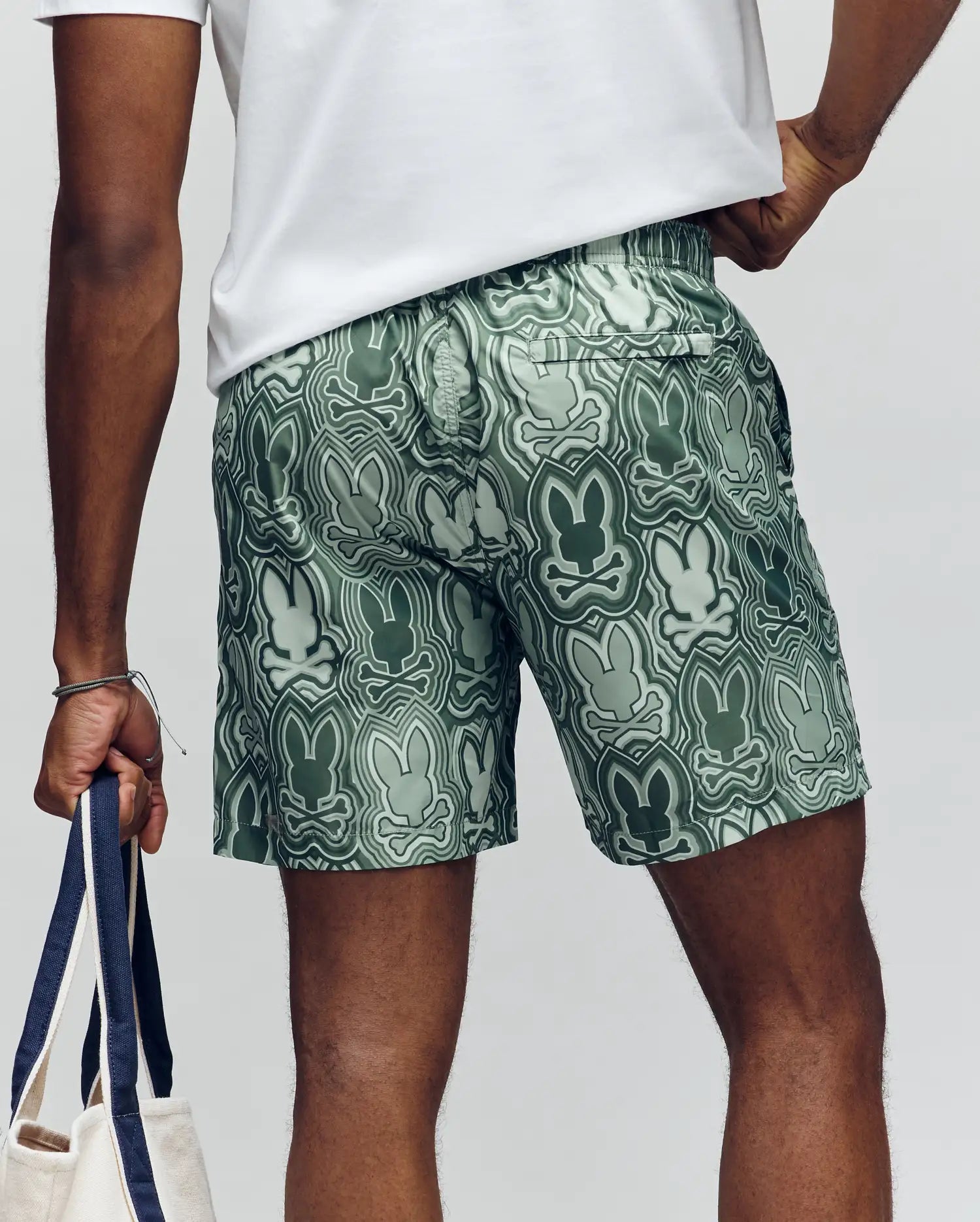 A person seen from behind wears Psycho Bunny MENS CHUCK ALL OVER PRINT LIGHTWEIGHT SWIM TRUNK (MB1000003)—green eco-friendly shorts with abstract bunny patterns—holding a tote in their left hand, emphasizing the sustainable design.