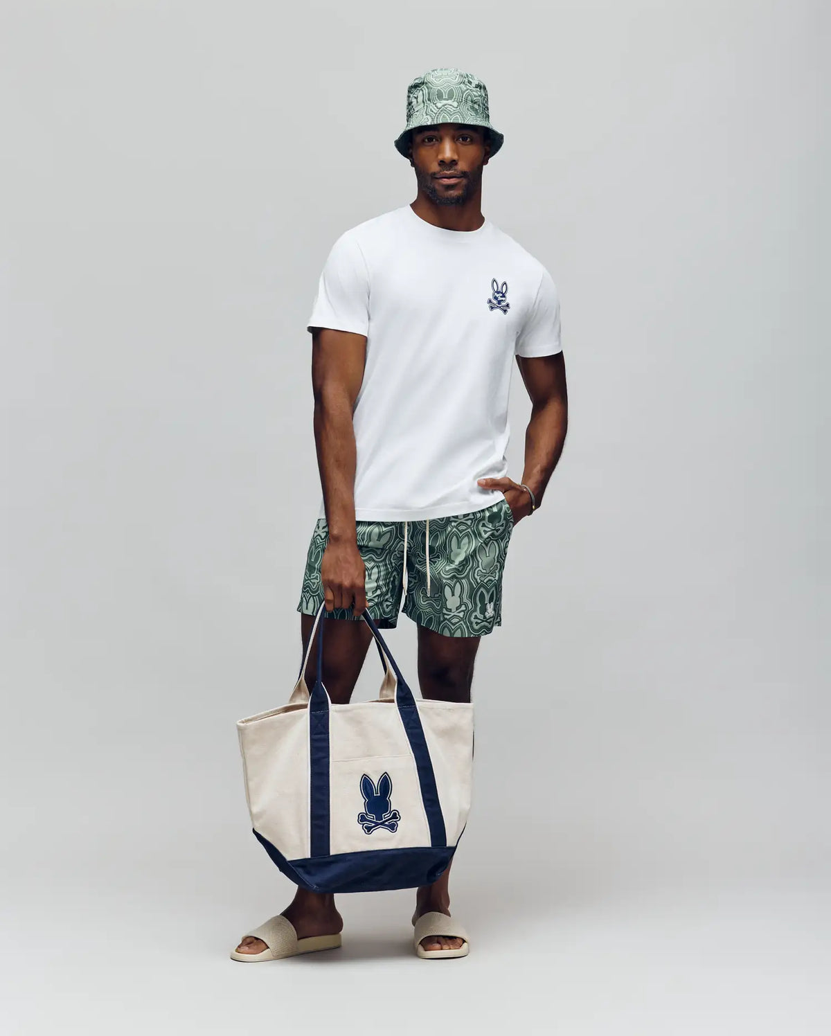 A man stands against a plain background wearing a white t-shirt, Psycho Bunny MENS CHUCK ALL OVER PRINT LIGHTWEIGHT SWIM TRUNK in green recycled polyester, a matching bucket hat, beige slides, and holds a beige and navy tote bag with the blue Psycho Bunny logo.