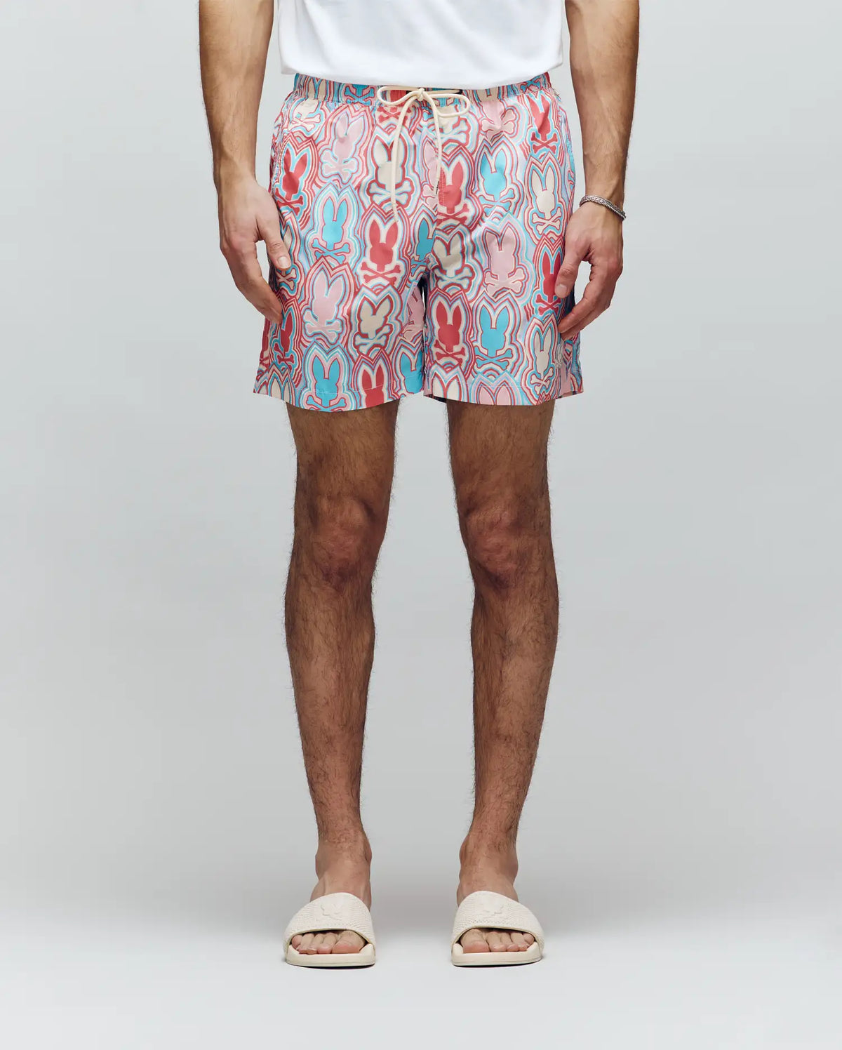 A person wearing a white T-shirt, white slide sandals, a bracelet, and Psycho Bunny MENS CHUCK ALL OVER PRINT LIGHTWEIGHT SWIM TRUNK - MB1000003 featuring colorful eco-conscious patterns, stands against a plain light background.