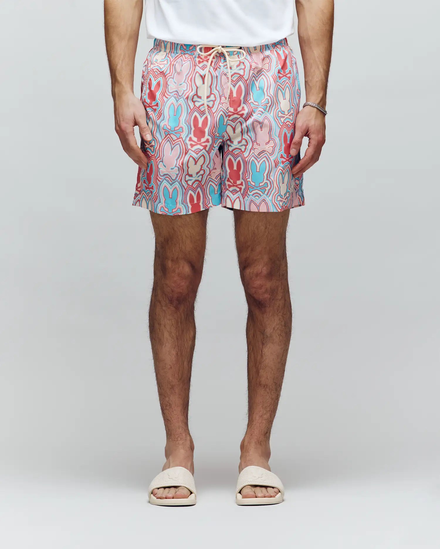 A person wearing a white T-shirt, white slide sandals, a bracelet, and Psycho Bunny MENS CHUCK ALL OVER PRINT LIGHTWEIGHT SWIM TRUNK - MB1000003 featuring colorful eco-conscious patterns, stands against a plain light background.