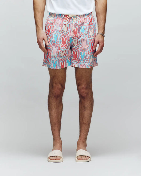 A person wearing a white T-shirt, white slide sandals, a bracelet, and Psycho Bunny MENS CHUCK ALL OVER PRINT LIGHTWEIGHT SWIM TRUNK - MB1000003 featuring colorful eco-conscious patterns, stands against a plain light background.