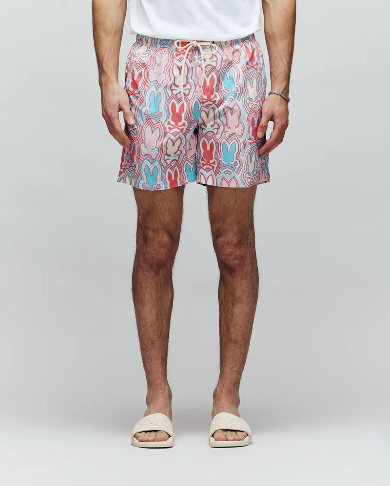 A person wearing a white T-shirt, white slide sandals, a bracelet, and Psycho Bunny MENS CHUCK ALL OVER PRINT LIGHTWEIGHT SWIM TRUNK - MB1000003 featuring colorful eco-conscious patterns, stands against a plain light background.