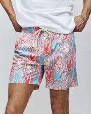 A person wears Psycho Bunny MENS CHUCK ALL OVER PRINT LIGHTWEIGHT SWIM TRUNK (MB1000003) featuring a pink, blue, red, and white bunny pattern on recycled polyester. Their hands are on their hips; a white shirt is partially visible.