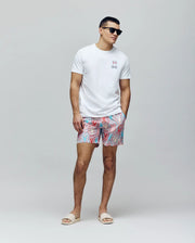 A man in Psycho Bunny MENS CHUCK ALL OVER PRINT LIGHTWEIGHT SWIM TRUNK - MB1000003 with blue, pink, and white patterns, a white tee featuring a small colorful bunny graphic, and black sunglasses stands against a plain light background.