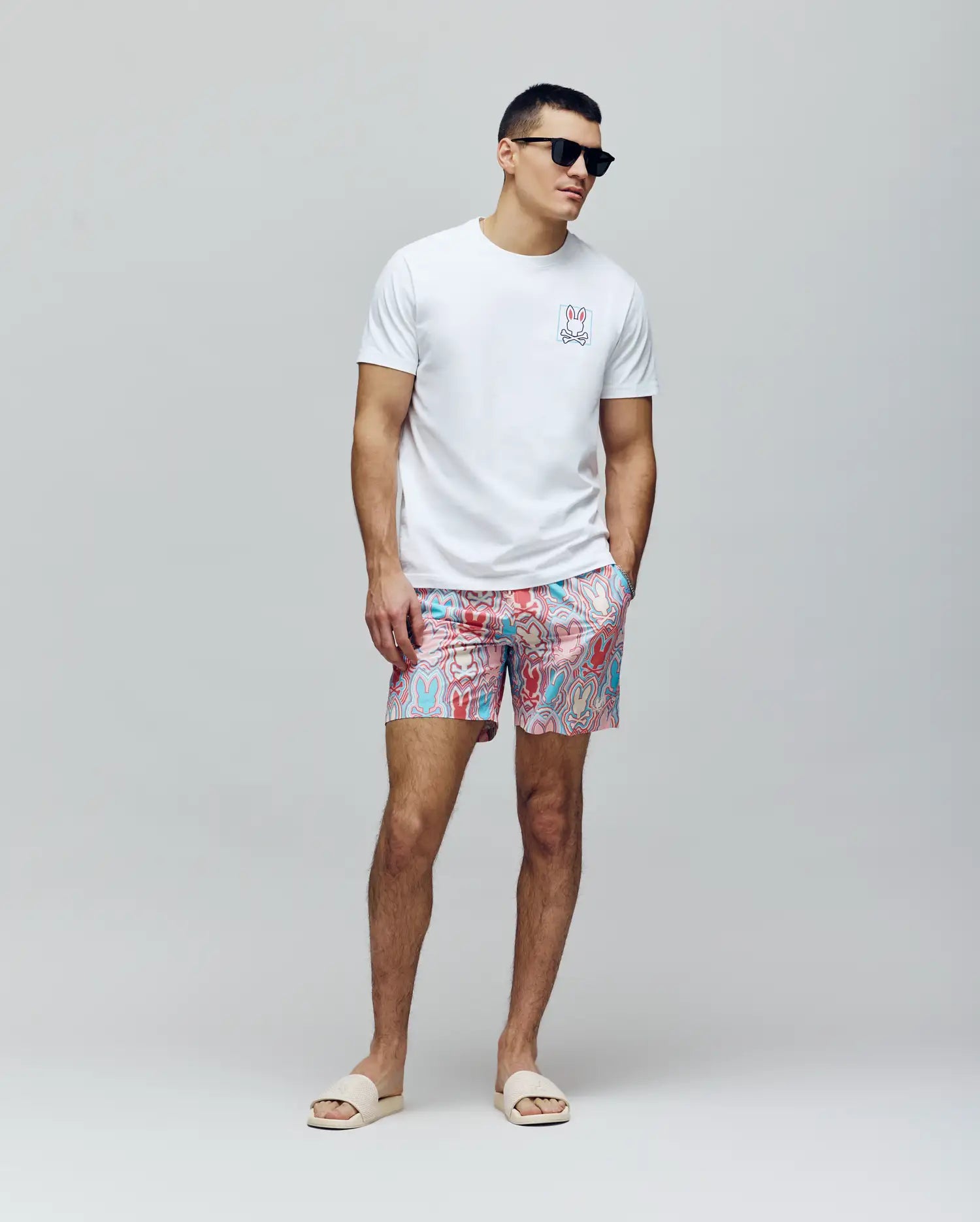 A man in Psycho Bunny MENS CHUCK ALL OVER PRINT LIGHTWEIGHT SWIM TRUNK - MB1000003 with blue, pink, and white patterns, a white tee featuring a small colorful bunny graphic, and black sunglasses stands against a plain light background.