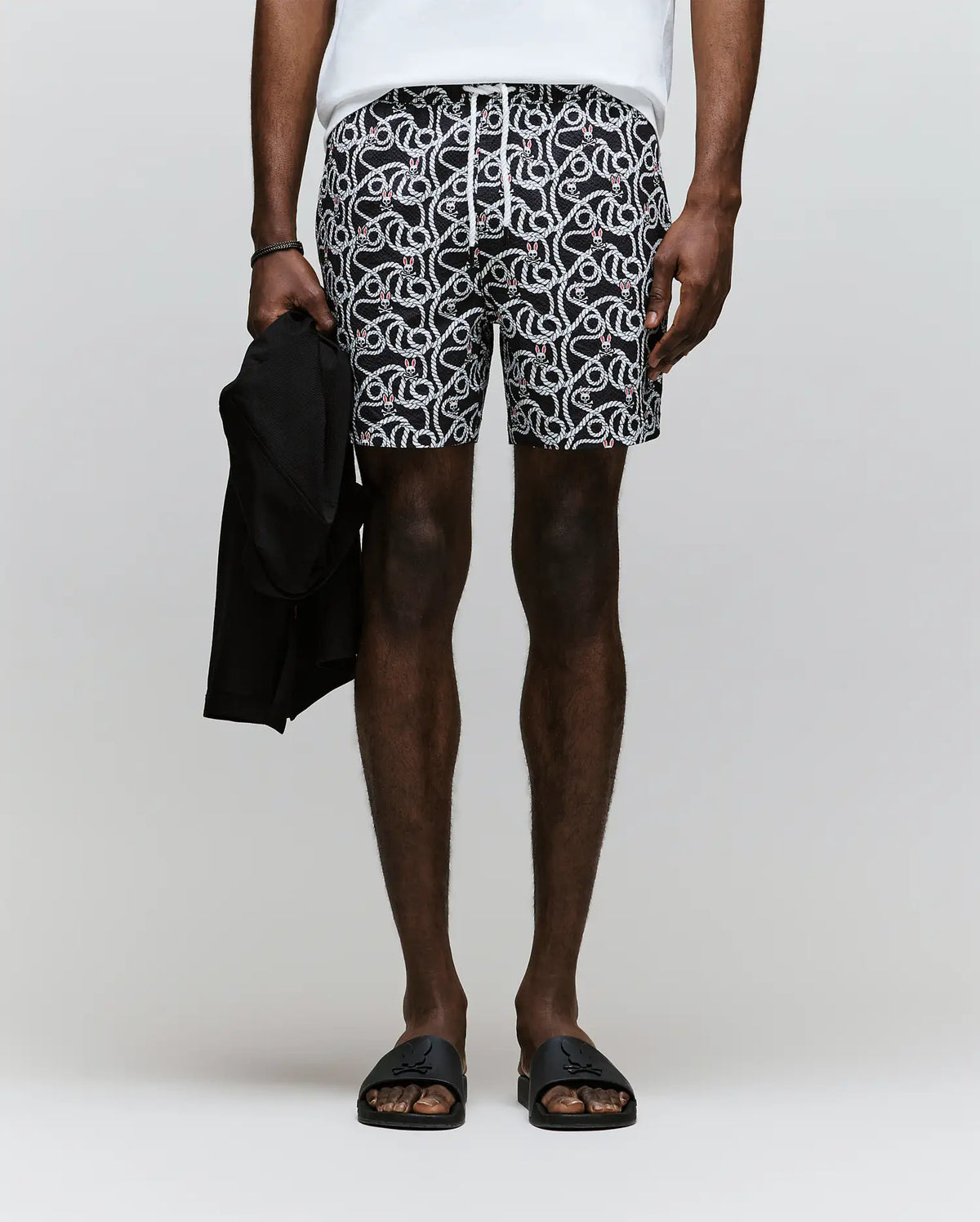 A person wears Psycho Bunny MENS CYRUS ALL OVER PRINT SWIM TRUNK - MB1000004 (black and white pattern), a white shirt, black sandals, and holds a black garment. The plain light background does not reveal the person’s face.