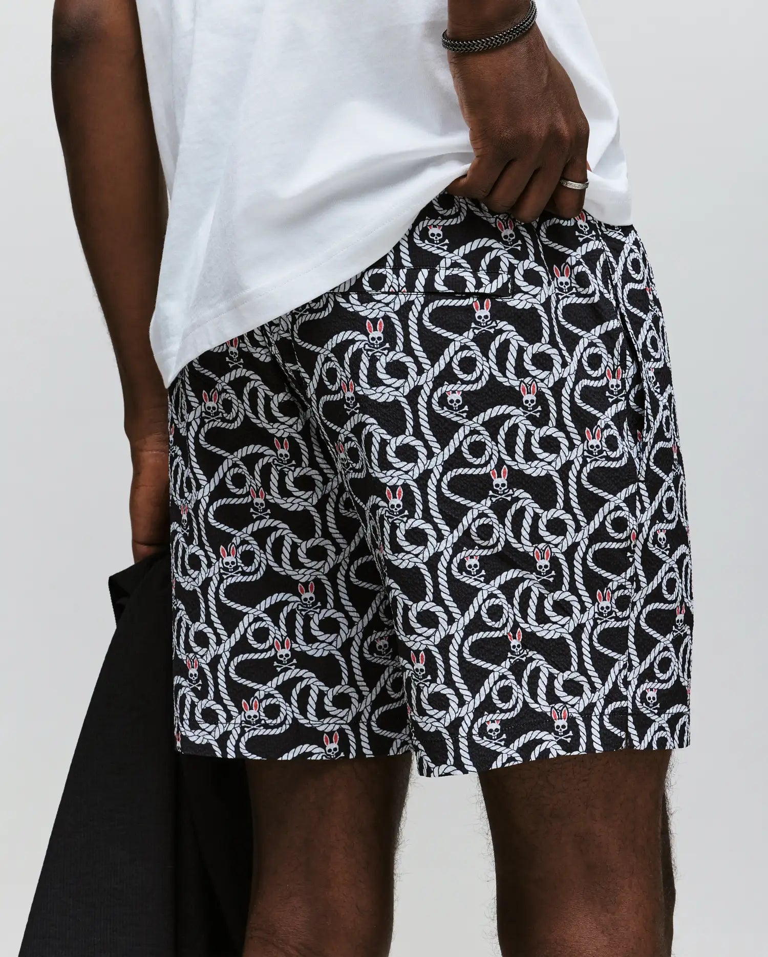 A man wears Psycho Bunny's MENS CYRUS ALL OVER PRINT SWIM TRUNK - MB1000004, featuring a red heart pattern and white rope design, paired with a white T-shirt and dark jacket; only his lower torso and upper legs are shown.