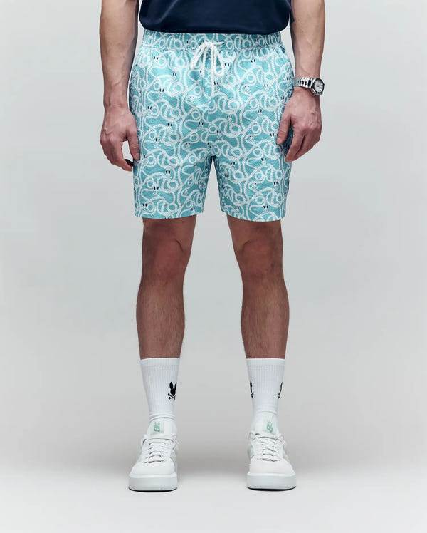 A person stands against a plain background, wearing a navy shirt, Psycho Bunny MENS CYRUS ALL OVER PRINT SWIM TRUNK - MB1000004, white socks with black logos, white sneakers, and a wristwatch.