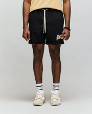 A person stands against a light gray background, wearing Psycho Bunny's MENS ELIAS SWIM TRUNK - MB1000008 (classic fit, yellow with white text), a yellow shirt, white socks with black stripes, and white sneakers. Photo is cropped below the shoulders.