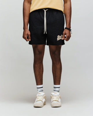 A person stands against a light gray background, wearing Psycho Bunny's MENS ELIAS SWIM TRUNK - MB1000008 (classic fit, yellow with white text), a yellow shirt, white socks with black stripes, and white sneakers. Photo is cropped below the shoulders.