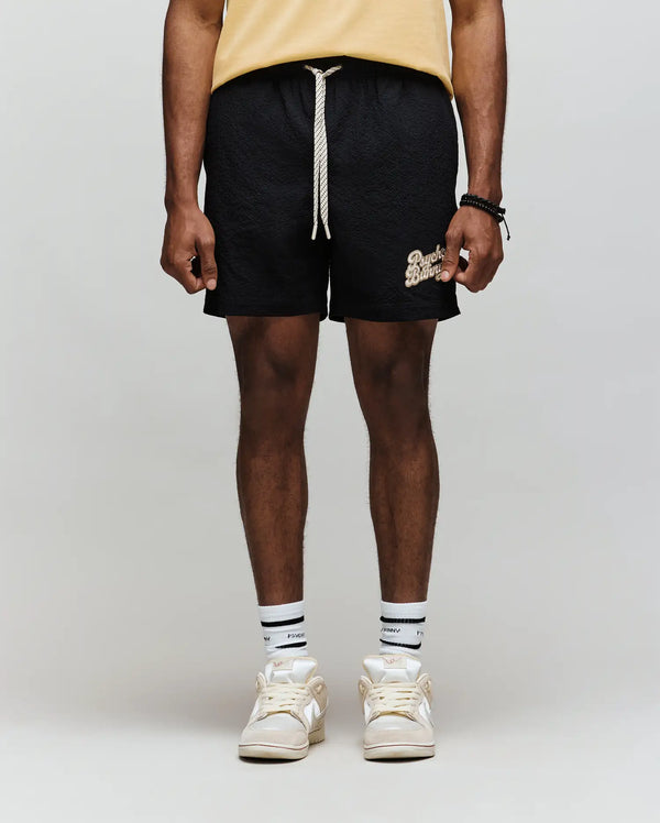 A person stands against a light gray background, wearing Psycho Bunny's MENS ELIAS SWIM TRUNK - MB1000008 (classic fit, yellow with white text), a yellow shirt, white socks with black stripes, and white sneakers. Photo is cropped below the shoulders.