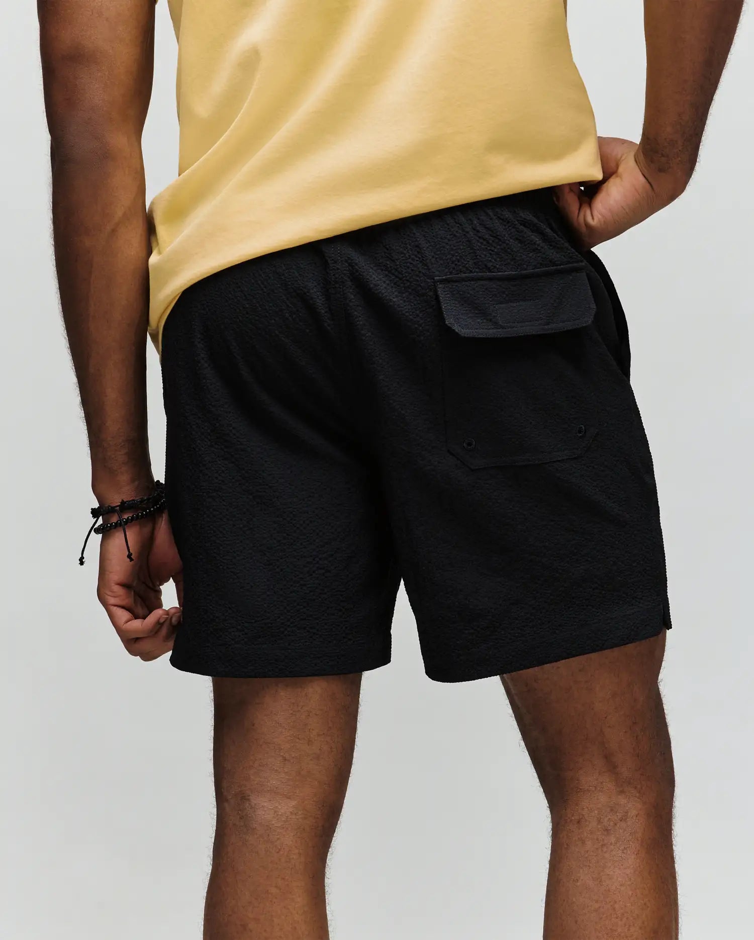 A person in a yellow shirt and Psycho Bunny MENS ELIAS SWIM TRUNK - MB1000008, classic fit and black color, stands with their back to the camera, showing a right pocket flap. Their left hand is on their hip; they wear black bracelets.