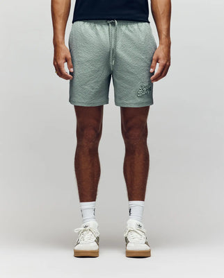 A person stands facing forward, showcasing only their lower half against a neutral background, wearing Psycho Bunny MENS ELIAS SWIM TRUNK - MB1000008 in a classic fit, styled with a black shirt, white crew socks, and white sneakers.