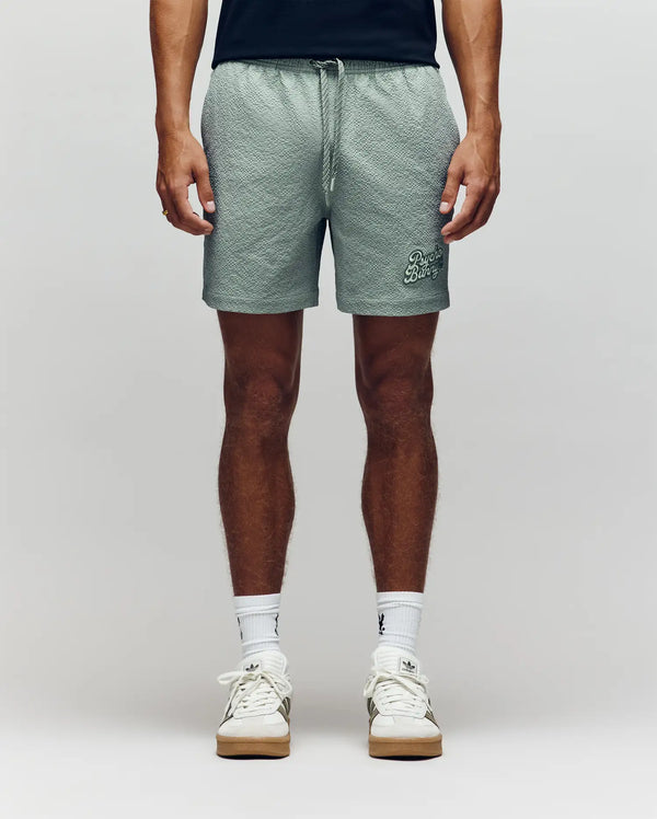 A person stands facing forward, showcasing only their lower half against a neutral background, wearing Psycho Bunny MENS ELIAS SWIM TRUNK - MB1000008 in a classic fit, styled with a black shirt, white crew socks, and white sneakers.