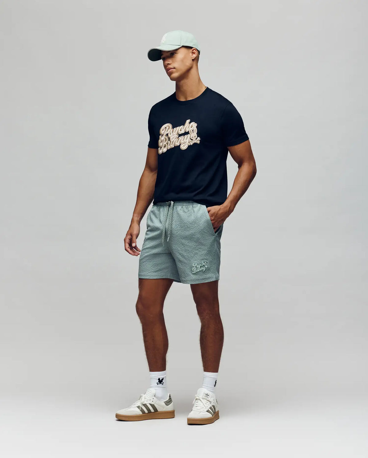 A man in a light green cap, black graphic t-shirt, Psycho Bunny MENS ELIAS SWIM TRUNK - MB1000008 with classic fit and no-chafe netting, white socks, and white sneakers stands against a plain light background.