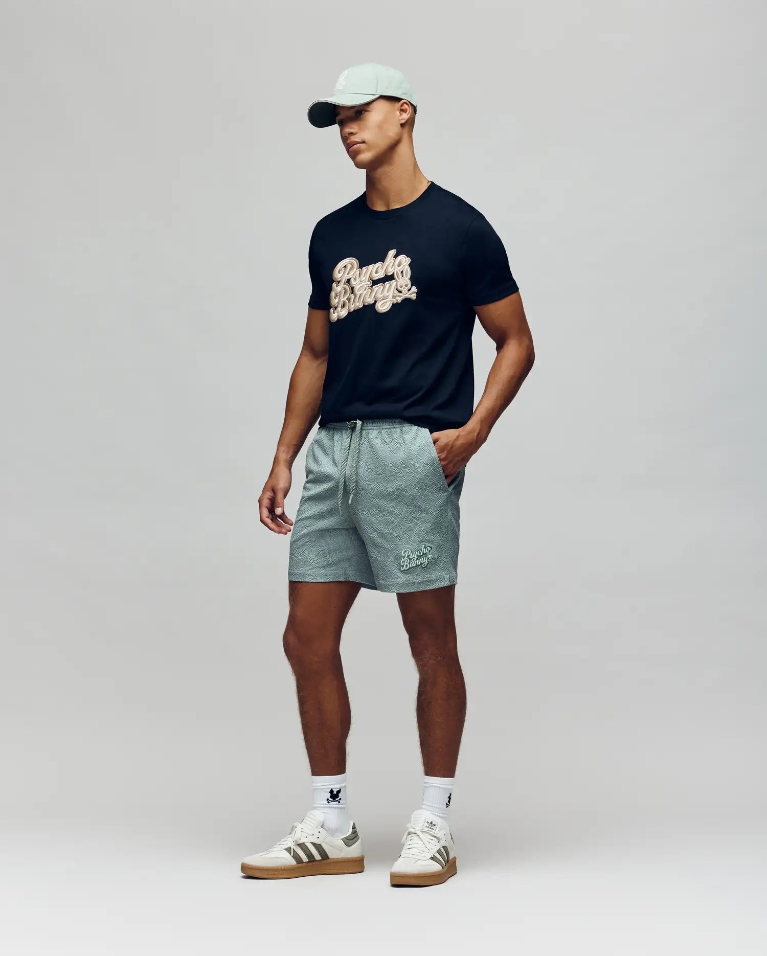 A man in a light green cap, black graphic t-shirt, Psycho Bunny MENS ELIAS SWIM TRUNK - MB1000008 with classic fit and no-chafe netting, white socks, and white sneakers stands against a plain light background.