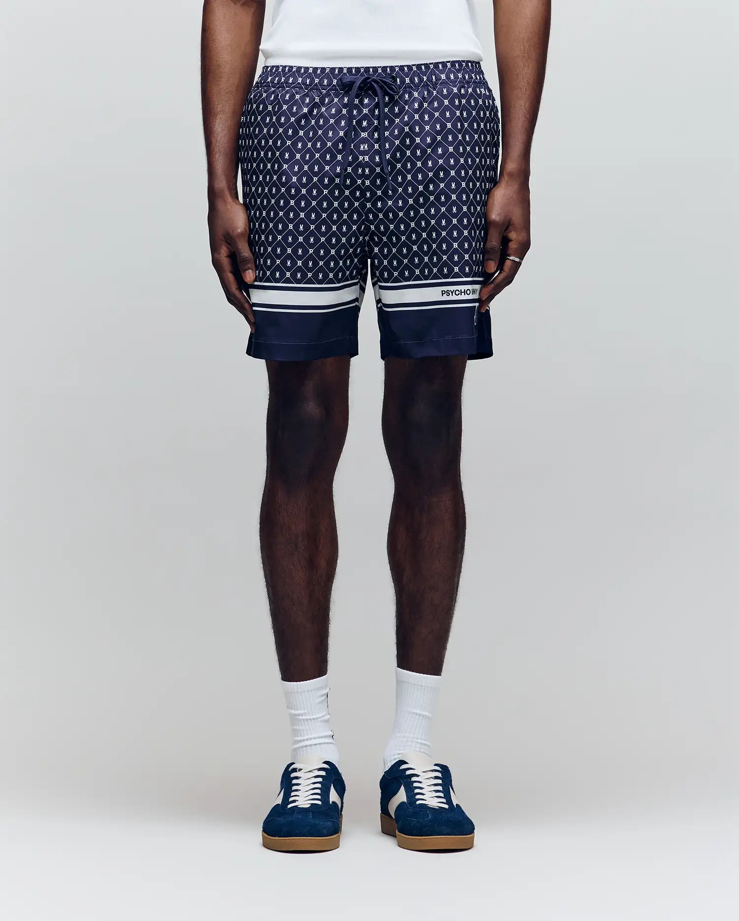 A man wears Psycho Bunny MENS HUNTER ALL OVER PRINT SWIM TRUNK (MB1000011), featuring a navy geometric pattern, quick-dry fabric, drawstring waist, white hem stripes, paired with white socks and blue sneakers against a plain light background.