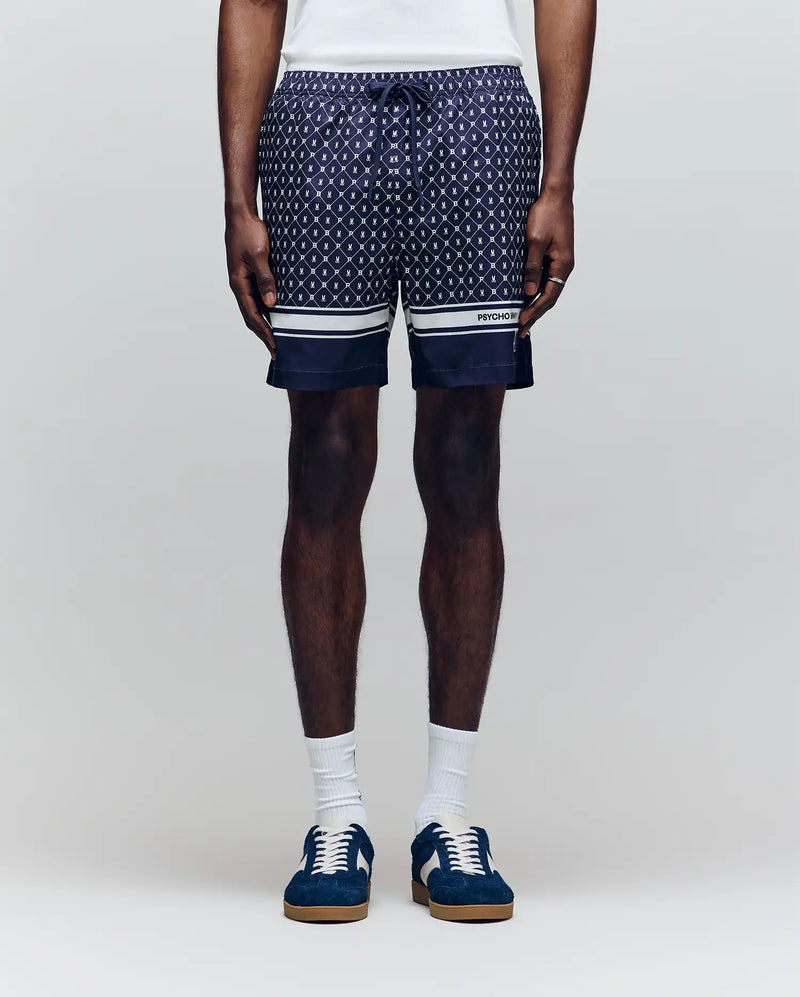 A man wears Psycho Bunny MENS HUNTER ALL OVER PRINT SWIM TRUNK (MB1000011), featuring a navy geometric pattern, quick-dry fabric, drawstring waist, white hem stripes, paired with white socks and blue sneakers against a plain light background.