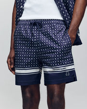 A person wearing Psycho Bunny MENS HUNTER ALL OVER PRINT SWIM TRUNK - MB1000011 in navy blue with a bunny pattern, white hem stripes, "PSYCHO BUNNY" branding on the left leg, and one hand in their pocket.