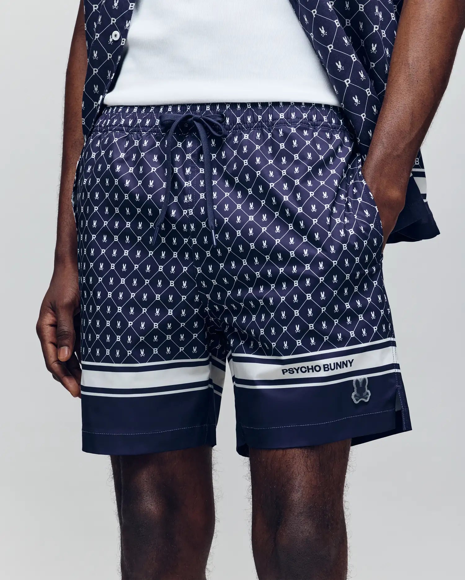 A person wearing Psycho Bunny MENS HUNTER ALL OVER PRINT SWIM TRUNK - MB1000011 in navy blue with a bunny pattern, white hem stripes, "PSYCHO BUNNY" branding on the left leg, and one hand in their pocket.