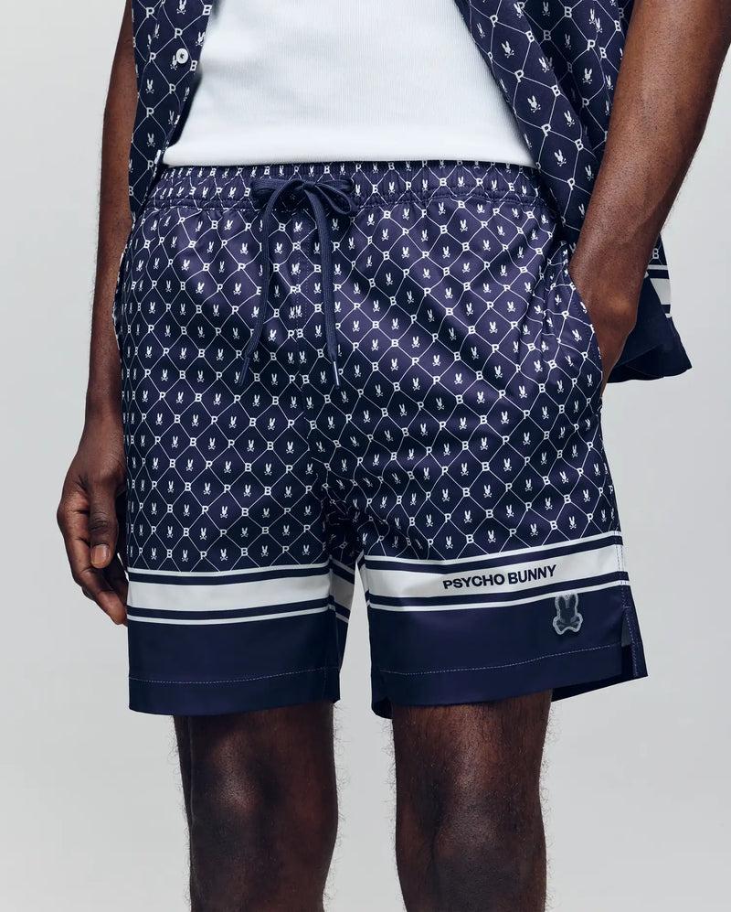 A person wearing Psycho Bunny MENS HUNTER ALL OVER PRINT SWIM TRUNK - MB1000011 in navy blue with a bunny pattern, white hem stripes, "PSYCHO BUNNY" branding on the left leg, and one hand in their pocket.