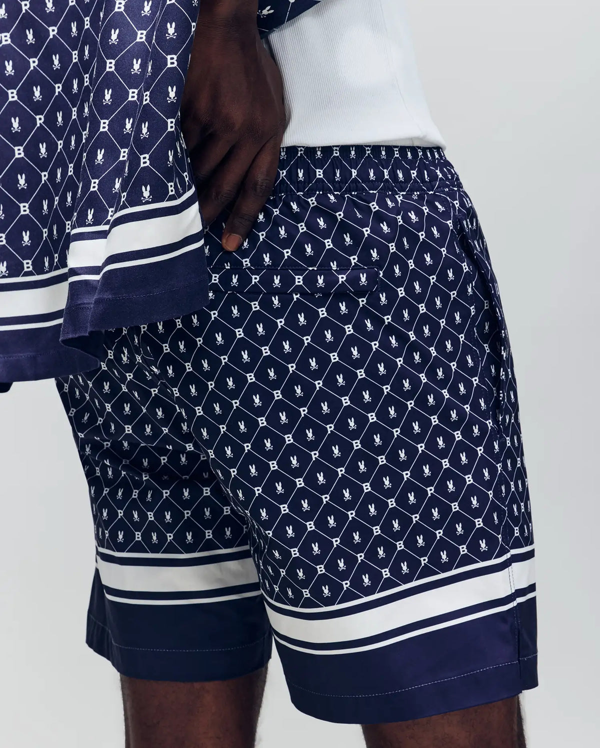 A person stands against a light gray background wearing Psycho Bunny's MENS HUNTER ALL OVER PRINT SWIM TRUNK (MB1000011), featuring a navy blue geometric and crest pattern, quick-dry fabric, and white horizontal stripes near the hems.