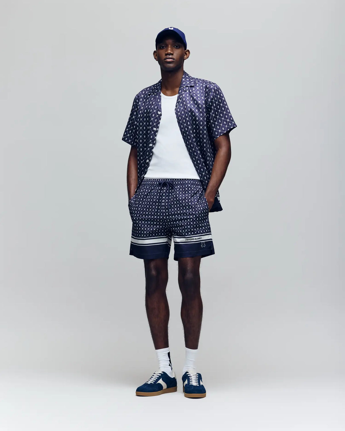 A man stands against a plain background wearing Psycho Bunny's MENS HUNTER ALL OVER PRINT SWIM TRUNK - MB1000011, a matching bunny pattern shirt, white tank top, navy baseball cap, blue and white sneakers, and white socks. His hands are in his shorts pockets.
