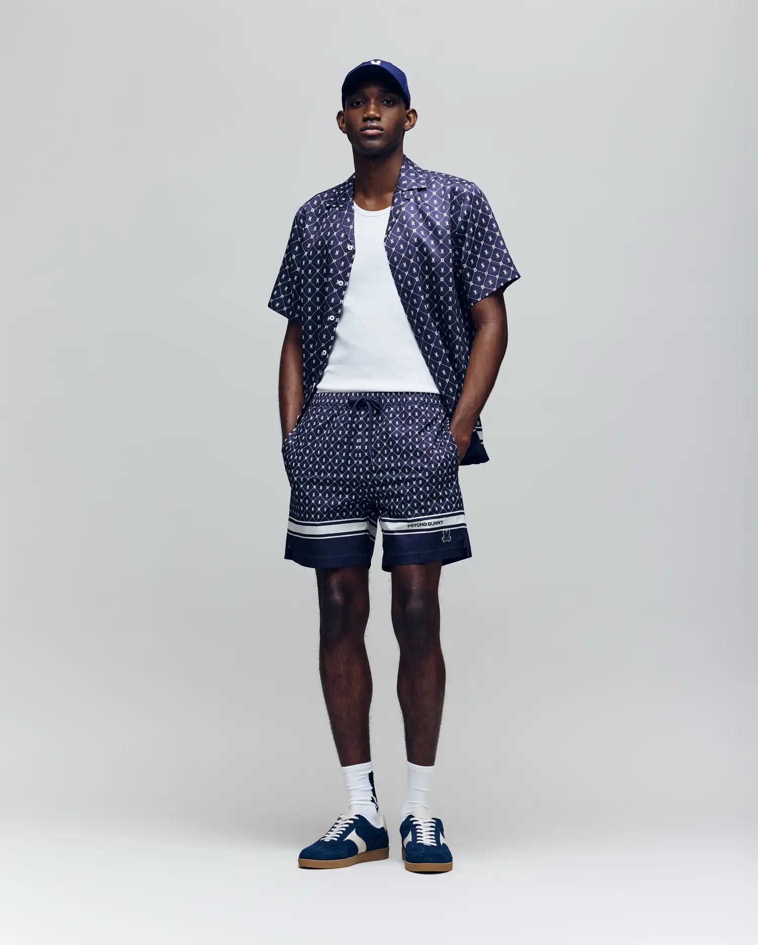 A man stands against a plain background wearing Psycho Bunny's MENS HUNTER ALL OVER PRINT SWIM TRUNK - MB1000011, a matching bunny pattern shirt, white tank top, navy baseball cap, blue and white sneakers, and white socks. His hands are in his shorts pockets.