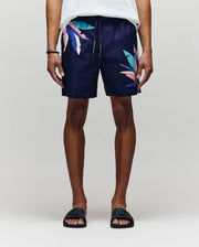 A person wears Psycho Bunny MENS SHANE SWIM TRUNK - MB1000029, featuring navy fabric with colorful abstract leaf patterns, paired with a white shirt and black slides, standing relaxed against a light gray background.