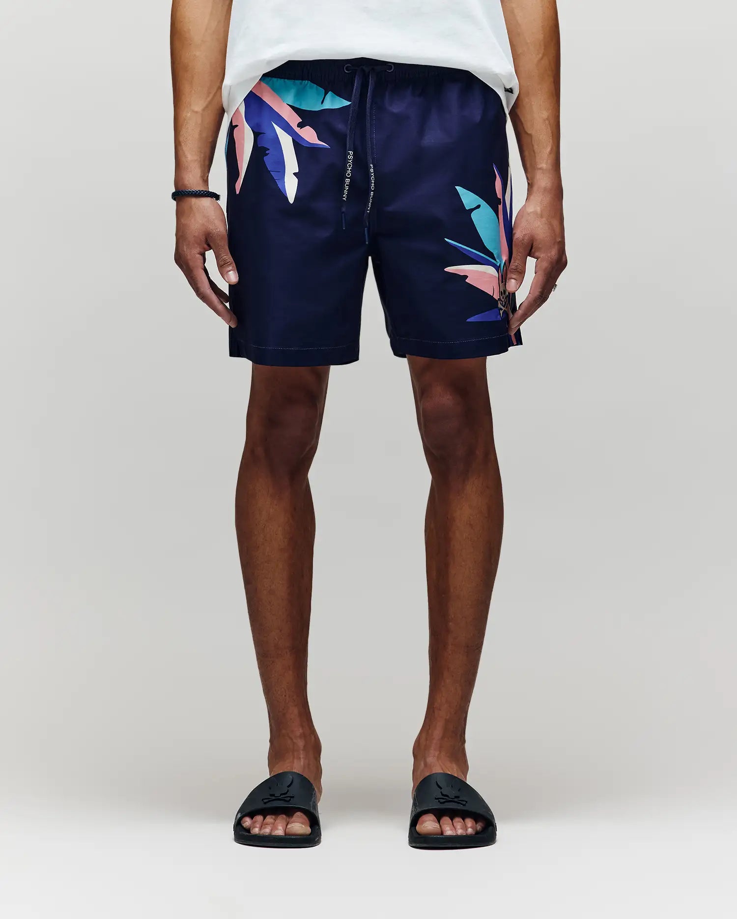 A person wears Psycho Bunny MENS SHANE SWIM TRUNK - MB1000029, featuring navy fabric with colorful abstract leaf patterns, paired with a white shirt and black slides, standing relaxed against a light gray background.