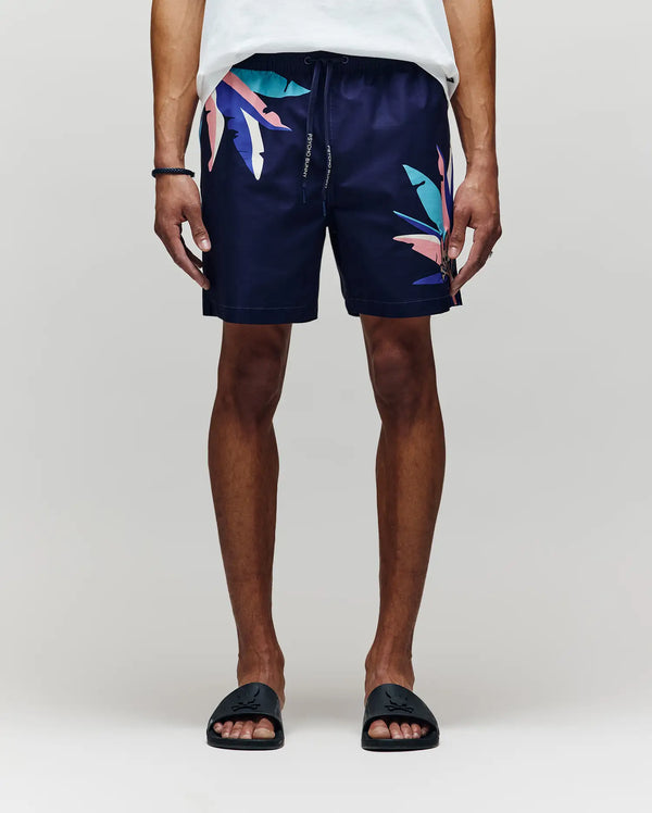 A person wears Psycho Bunny MENS SHANE SWIM TRUNK - MB1000029, featuring navy fabric with colorful abstract leaf patterns, paired with a white shirt and black slides, standing relaxed against a light gray background.