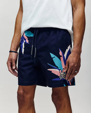 Wearing Psycho Bunny’s MENS SHANE SWIM TRUNK - MB1000029, featuring navy blue abstract leaf patterns and a small bunny logo, paired with a white tee, exudes relaxed luxury. Only the lower torso and upper legs are visible.