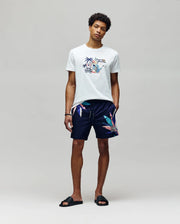 A man stands against a plain background wearing a white graphic tee, black slides, and Psycho Bunny MENS SHANE SWIM TRUNK - MB1000029 featuring a colorful leaf print, hands in pockets and exuding relaxed luxury.