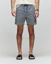 A person stands facing forward, wearing a cream shirt, black slide sandals, and Psycho Bunny MENS ZANE SWIM TRUNK - MB1000030—navy and white vertical striped swim shorts with a no-chafe net and drawstring. Hands are by their sides; background is light gray.