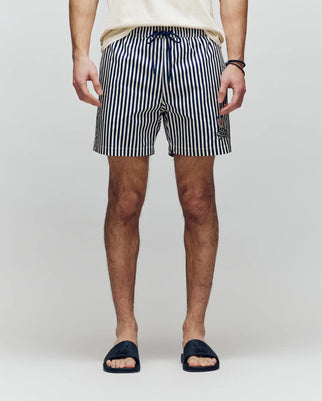 A person stands facing forward, wearing a cream shirt, black slide sandals, and Psycho Bunny MENS ZANE SWIM TRUNK - MB1000030—navy and white vertical striped swim shorts with a no-chafe net and drawstring. Hands are by their sides; background is light gray.
