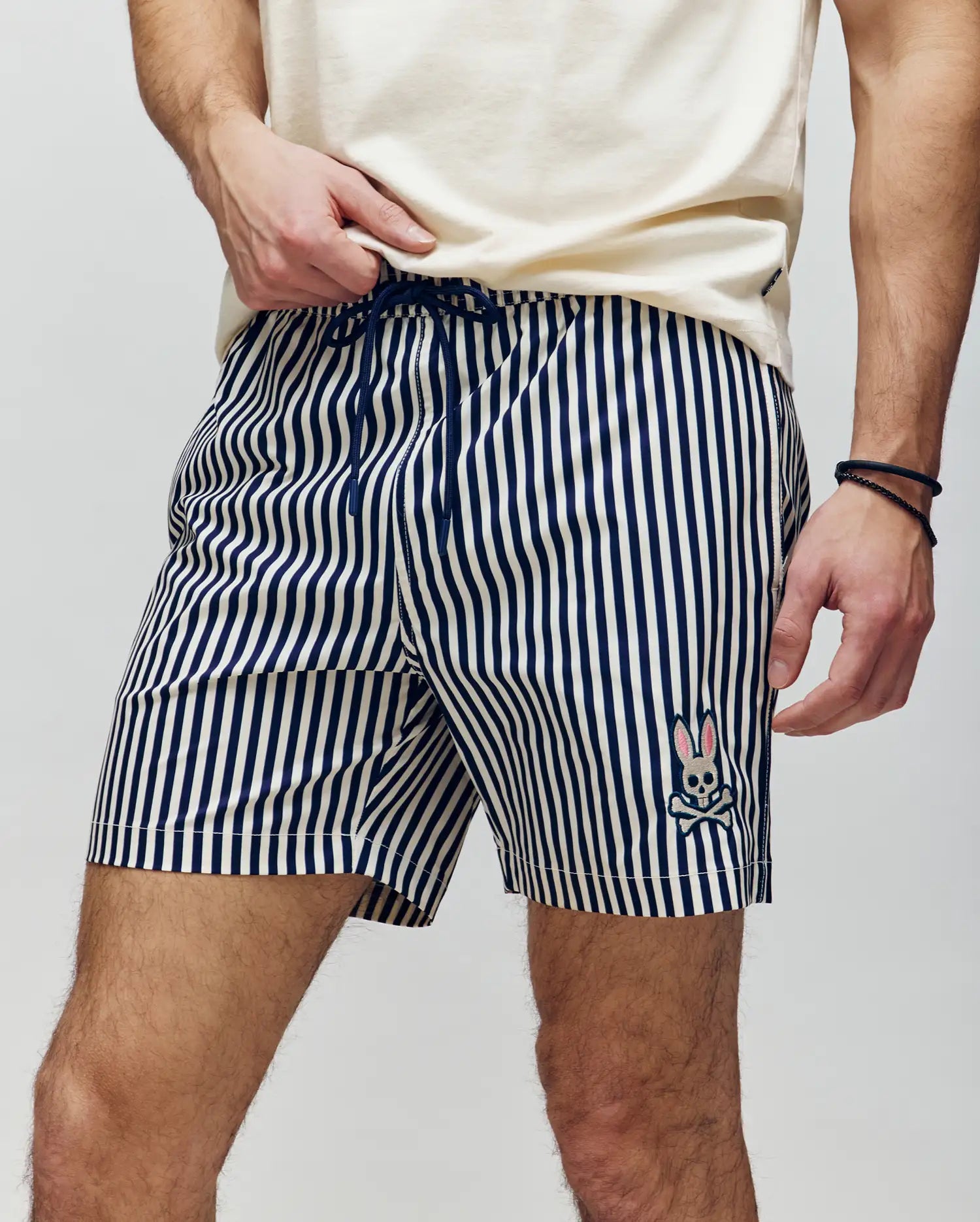 A person in Psycho Bunny MENS ZANE SWIM TRUNK - MB1000030, featuring navy and white vertical stripes and a bunny face logo on the left leg, stands against a plain background wearing a light shirt and holding a waterproof bag.