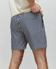 A person is shown from the back wearing Psycho Bunny’s MENS ZANE SWIM TRUNK - MB1000030, featuring blue and white vertical stripes, a no-chafe net, and a light-colored shirt, with one hand in their pocket.