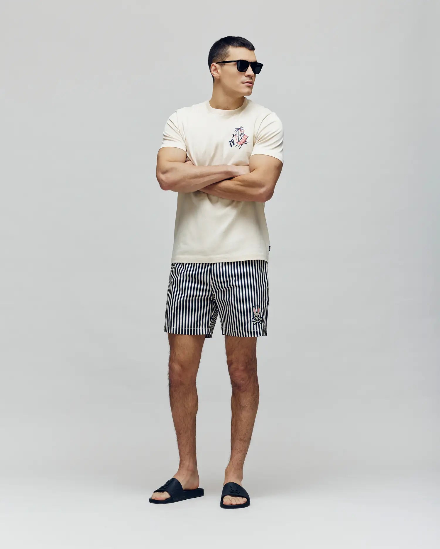 A man stands with arms crossed, wearing sunglasses, a white tee, black sandals, and Psycho Bunny MENS ZANE SWIM TRUNK - MB1000030 featuring black and white vertical stripes, against a plain light background.