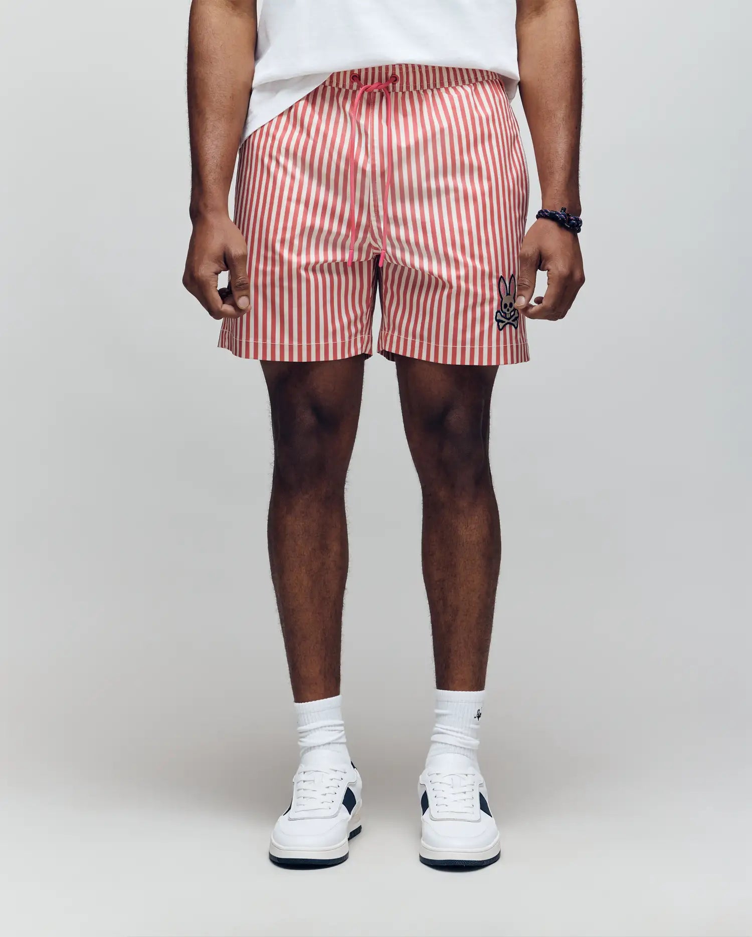 A person stands against a plain background wearing Psycho Bunny MENS ZANE SWIM TRUNK - MB1000030, a white shirt, white crew socks, and white sneakers. The shorts have red and white vertical stripes and feature a small black bunny logo. Hands relaxed at their sides.