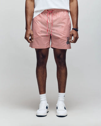 A person stands against a plain background wearing Psycho Bunny MENS ZANE SWIM TRUNK - MB1000030, a white shirt, white crew socks, and white sneakers. The shorts have red and white vertical stripes and feature a small black bunny logo. Hands relaxed at their sides.