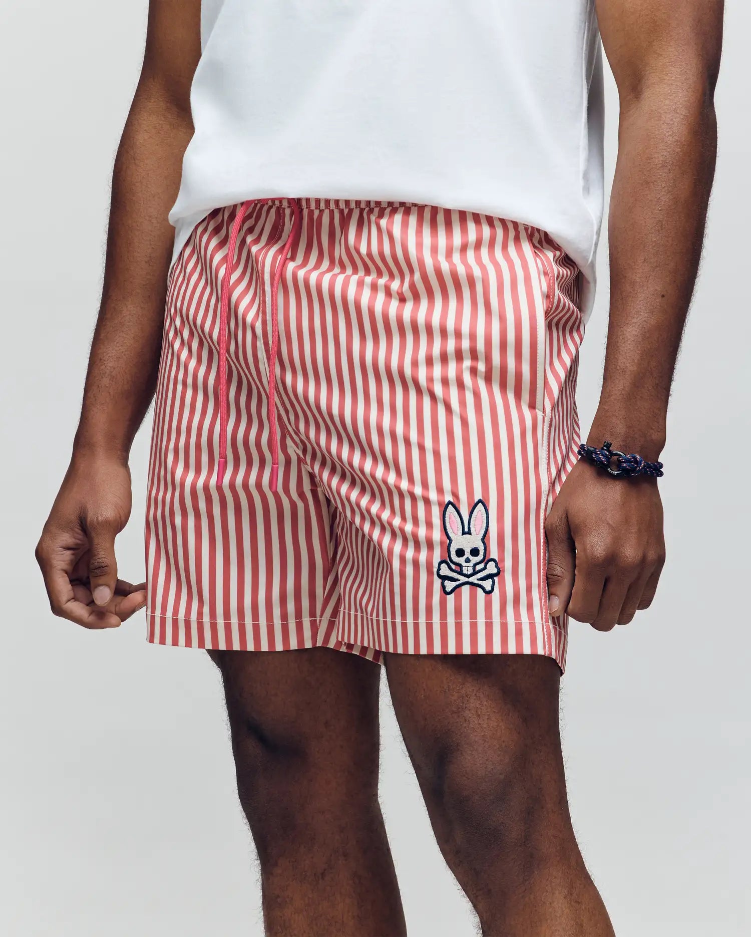 A man wears Psycho Bunny MENS ZANE SWIM TRUNK - MB1000030 featuring red and white vertical stripes, a cartoon bunny and crossbones patch, comfy no-chafe net lining, plus a white tee and dark bracelet. His hand rests casually by his side.