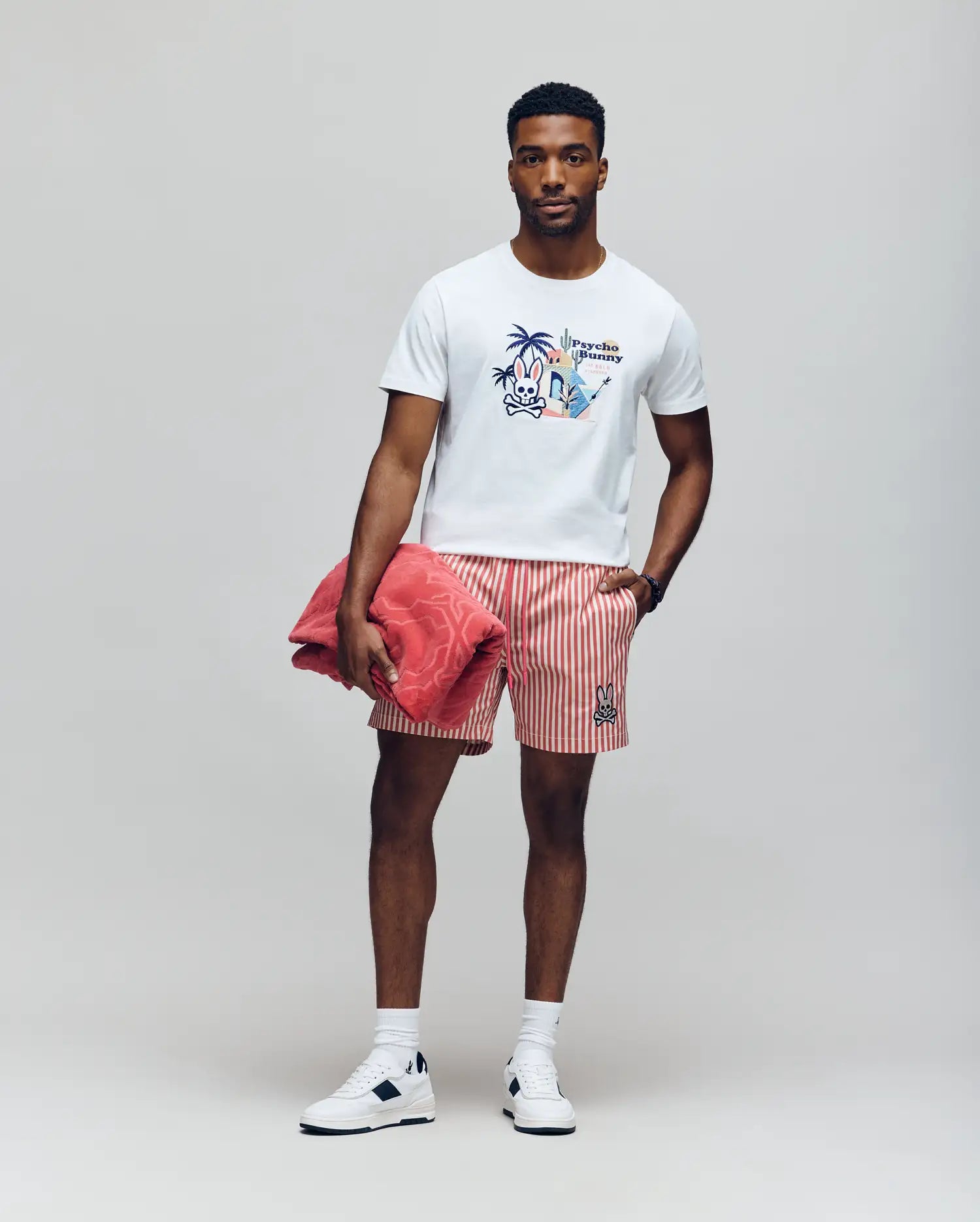 A man stands against a plain background wearing a white graphic T-shirt, Psycho Bunny MENS ZANE SWIM TRUNK - MB1000030 red and white striped shorts with no-chafe net, white socks, and sneakers. He holds a pink towel under one arm and has his other hand in his pocket.